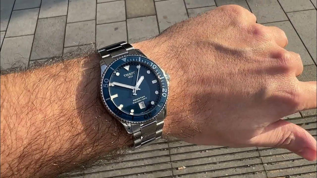 Hand wearing a Tissot watch with a blue face on a pavement background